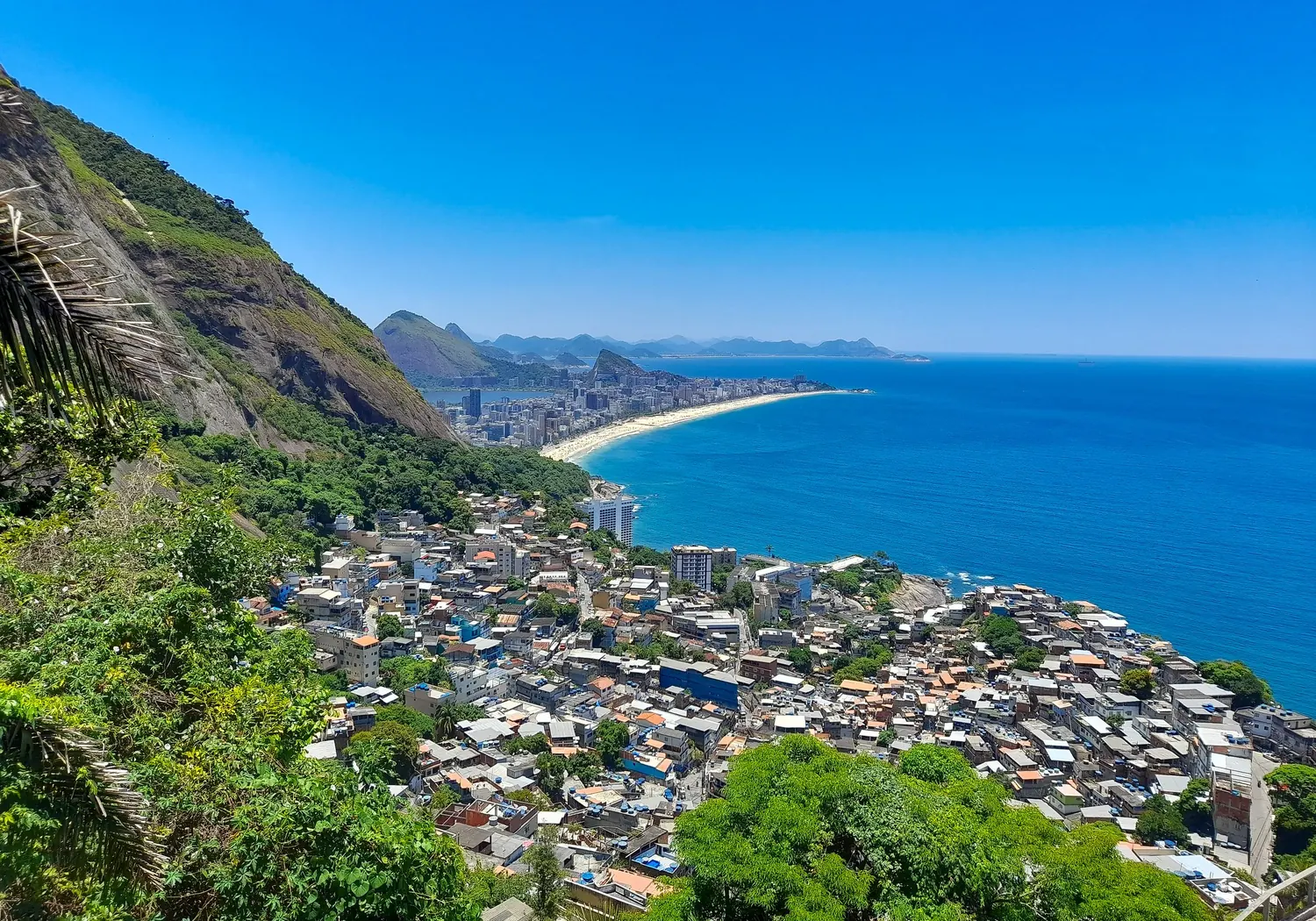 Vy över tätt byggda bostadsområden på en grön sluttning ned mot havet i Rio de Janeiro.